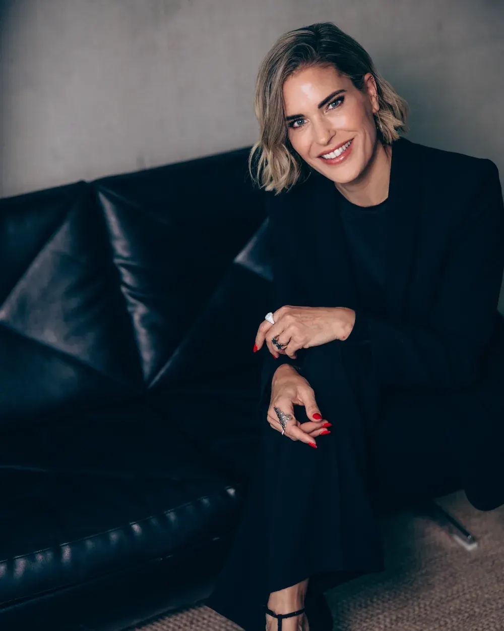 Elke Rock lacht in die Kamera, schwarzes Outfit auf Ledersofa